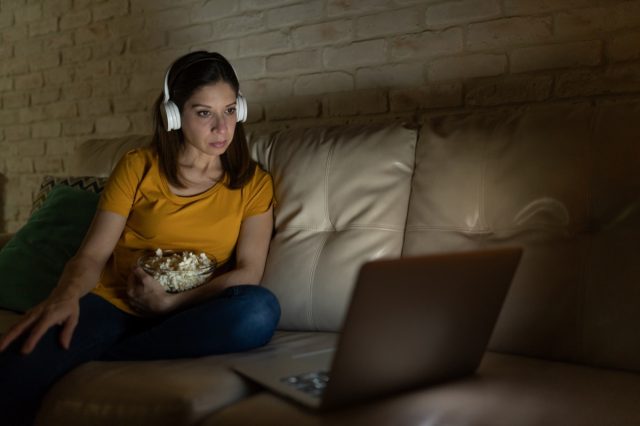 Una mujer de unos cuarenta años usa audífonos y come palomitas de maíz mientras ve una película en el servicio de transmisión en una computadora portátil por la noche.